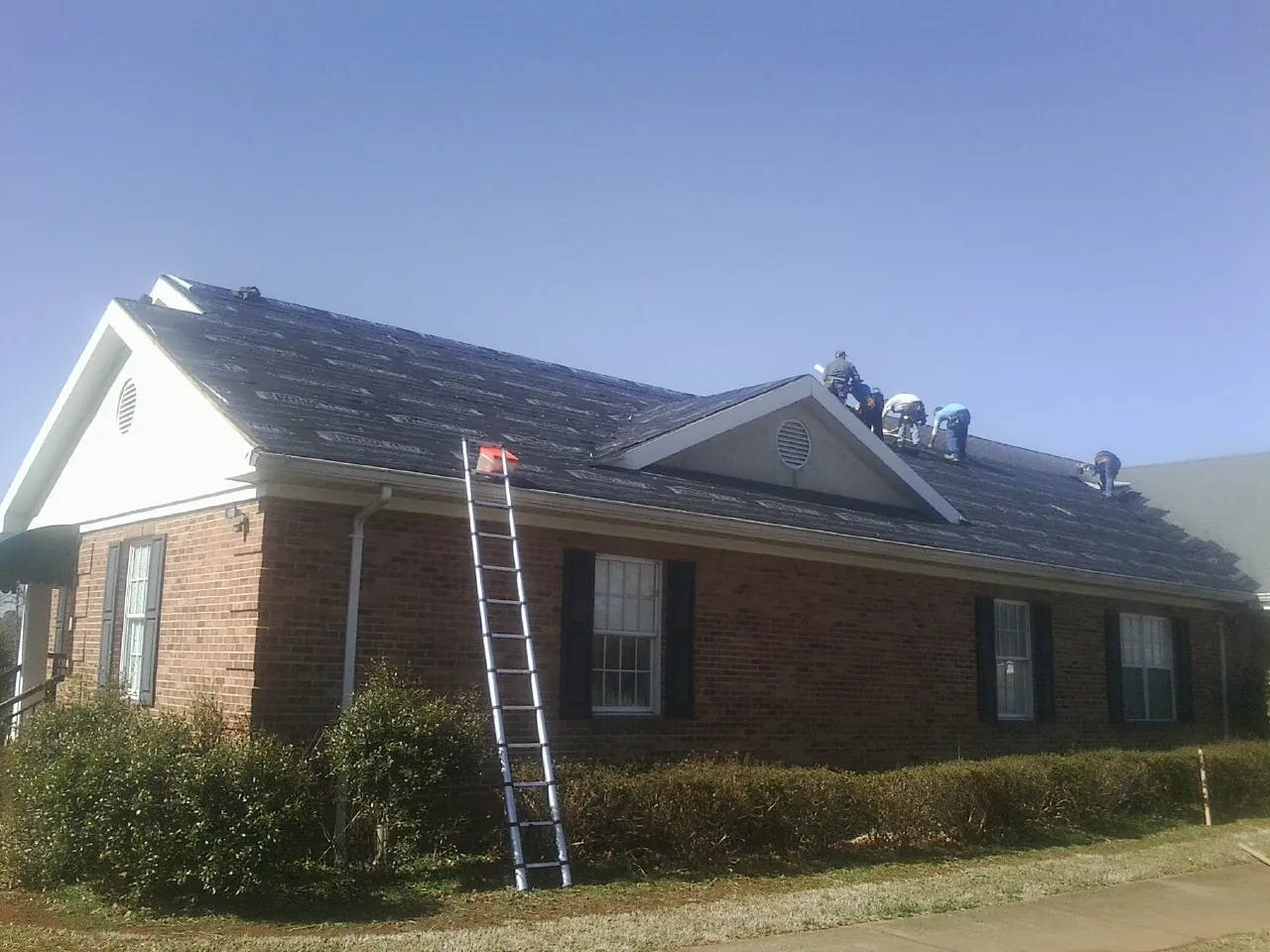 Crew tearing shingles off a brick ranch roof with a ladder set — Emergency Roof Repair work by our crew in High Bridge