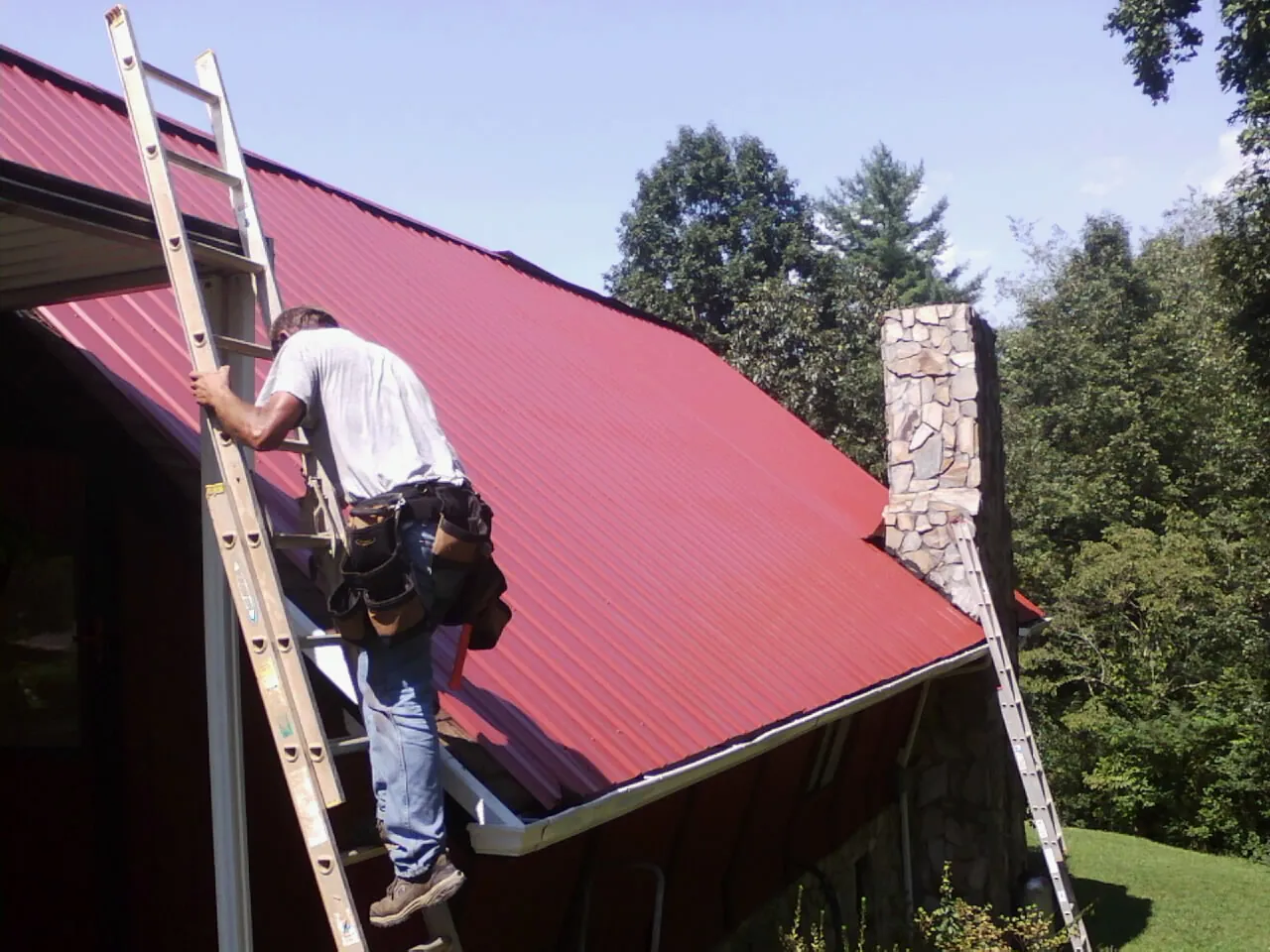 Roofer on a ladder beside a red metal roof and stone chimney — Soffit & Fascia Repair by Roofing Brothers in High Bridge